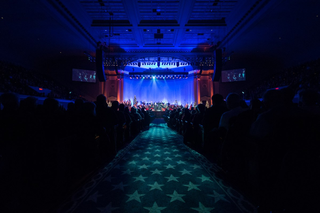 Ein großer Zuschauerraum mit Sitzplätzen, eine Bühne mit Musikern und Deckenleuchten und -fernseher, was auf ein Konzerthalle während der Nationalkonvention 2019 hindeutet.