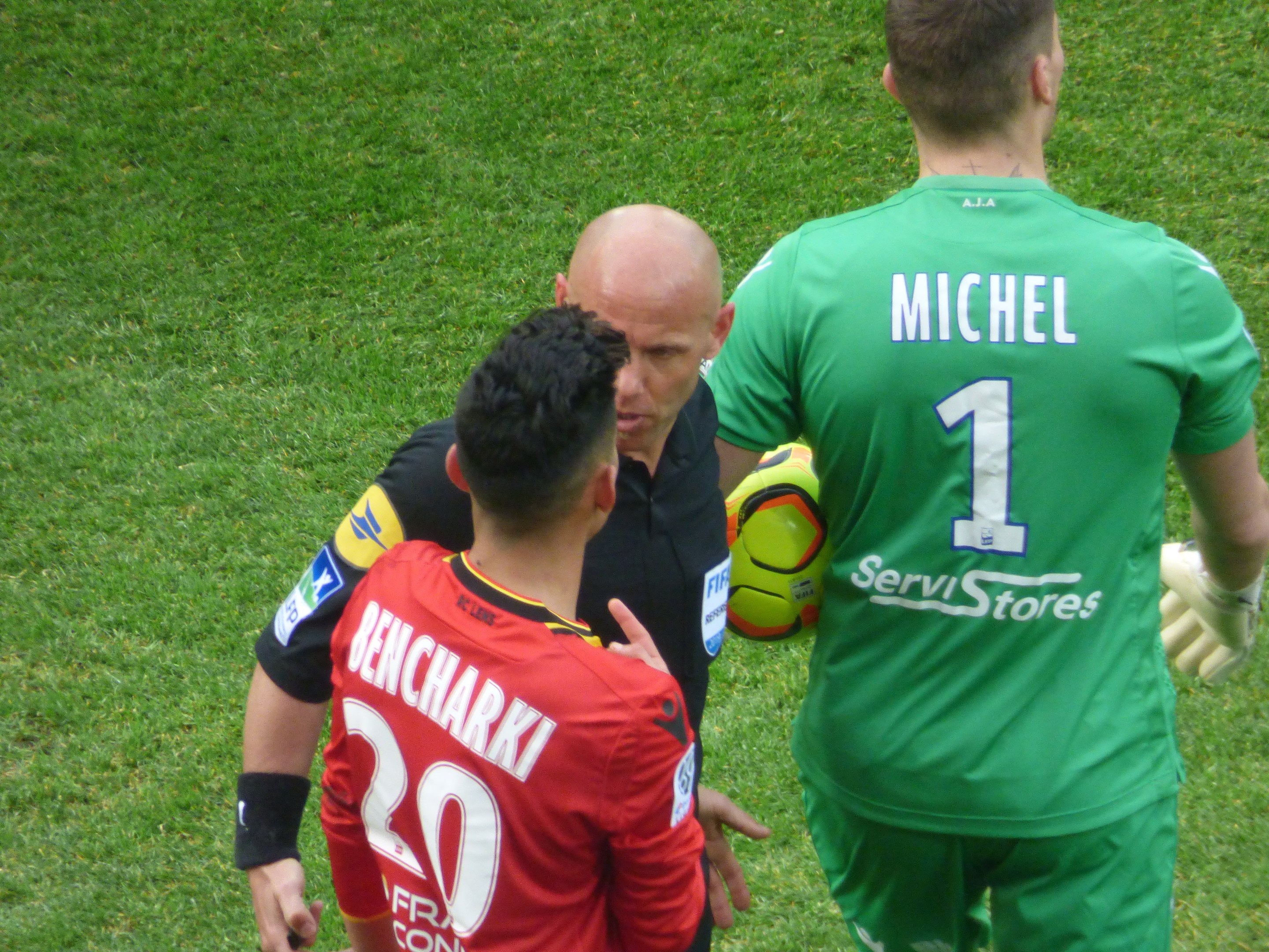 Schiedsrichter spricht mit zwei Fußballspielern auf einem grünen Rasen, einer hält den Ball.