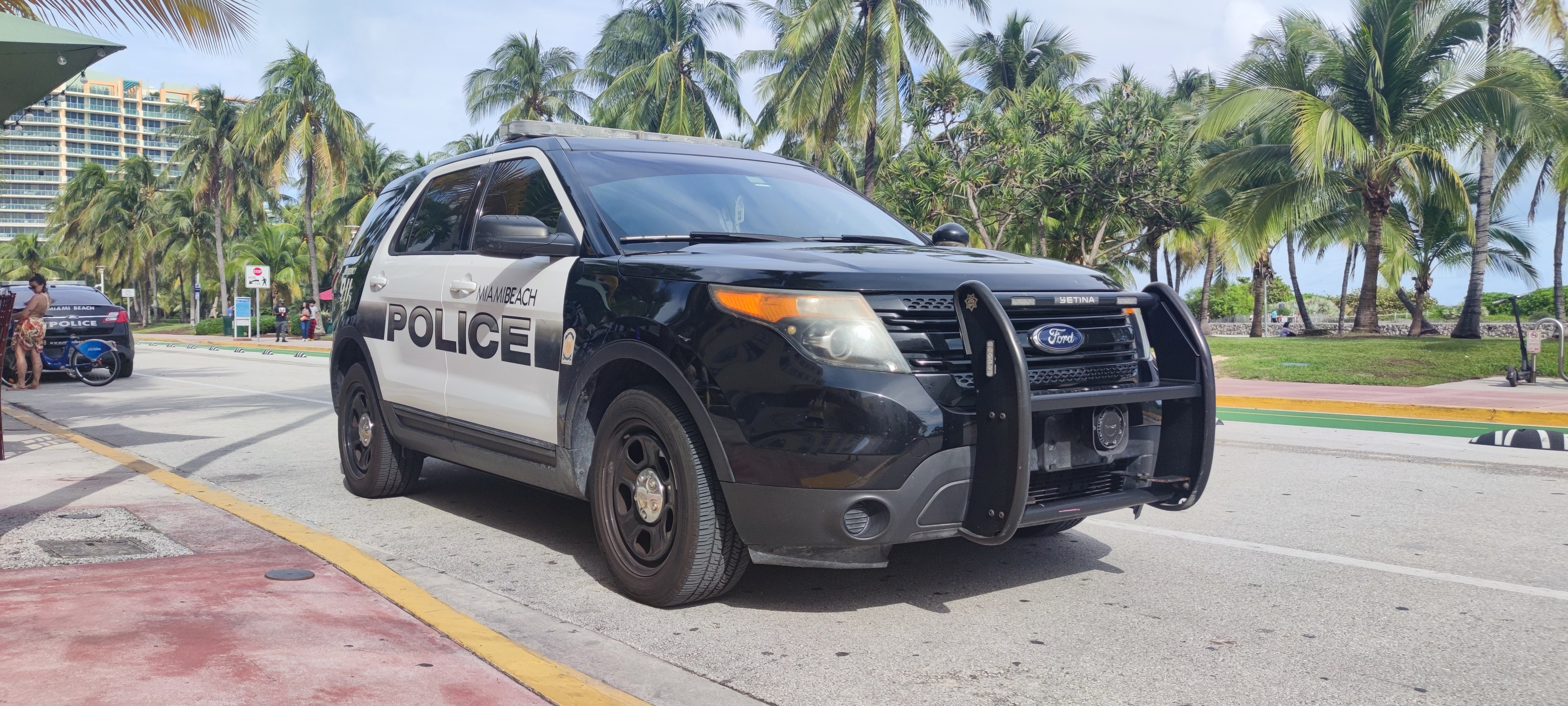 Polizeiauto auf dem Seitenstreifen geparkt mit Menschen, Fahrrädern, Gras, Bäumen, Gebäuden und einem klaren blauen Himmel im Hintergrund; das Vorderrad des Autos ist sichtbar.