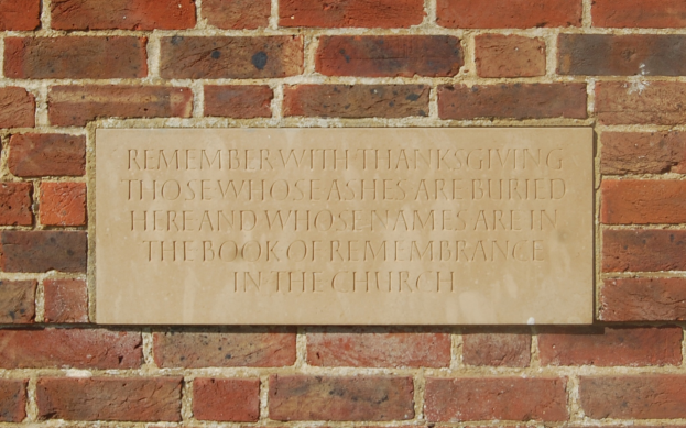 Eine Backsteinwand mit einer Tafel, auf der steht: "Erinnere dich mit Dankbarkeit an diejenigen, deren Asche hier begraben ist und deren Namen im Buch der Erinnerung in der Kirche stehen."