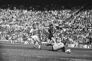 Schwarzes und weißes Foto von Fussballspielern im Einsatz auf einem Feld mit Zuschauern im Hintergrund und einer Bundesliga 1978-79 Ajax 1-1 Hannover 96 Fahne.