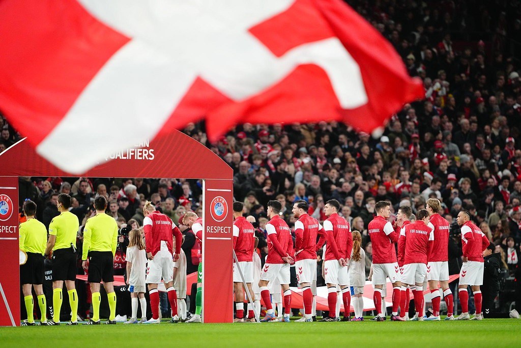 Eine Gruppe von Menschen auf einem Fuballfeld mit einer roten und weißen Flagge im Vordergrund, ein Bogen mit der Aufschrift "Bayern Munich vs Bayern Munich Betting Tips & Preview" im Hintergrund und eine große Menge im Stadion.