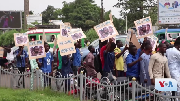 Eine Gruppe von Menschen mit Schildern, wahrscheinlich protestierend, steht vor einem Zaun mit Fahrzeugen, Bäumen, Pfählen, Absperrungen und einem Bildschirm im Hintergrund, unter einem sichtbaren Himmel mit Gras am unteren Bildrand.