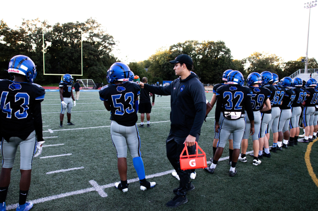 Football coach in a cap and players in helmets on a field with trees, poles, lights, railings, a goal post, and a clear blue sky.