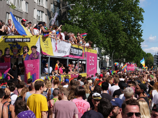 Eine große Menschenmenge marschiert auf der Straße während des Christopher Street Day 2019 in Berlin, viele tragen Mützen und Schutzbrillen, einige halten Fahnen, mit Bannern, Laternenmasten, Bäumen und Gebäuden im Hintergrund unter einem bewölkten Himmel.