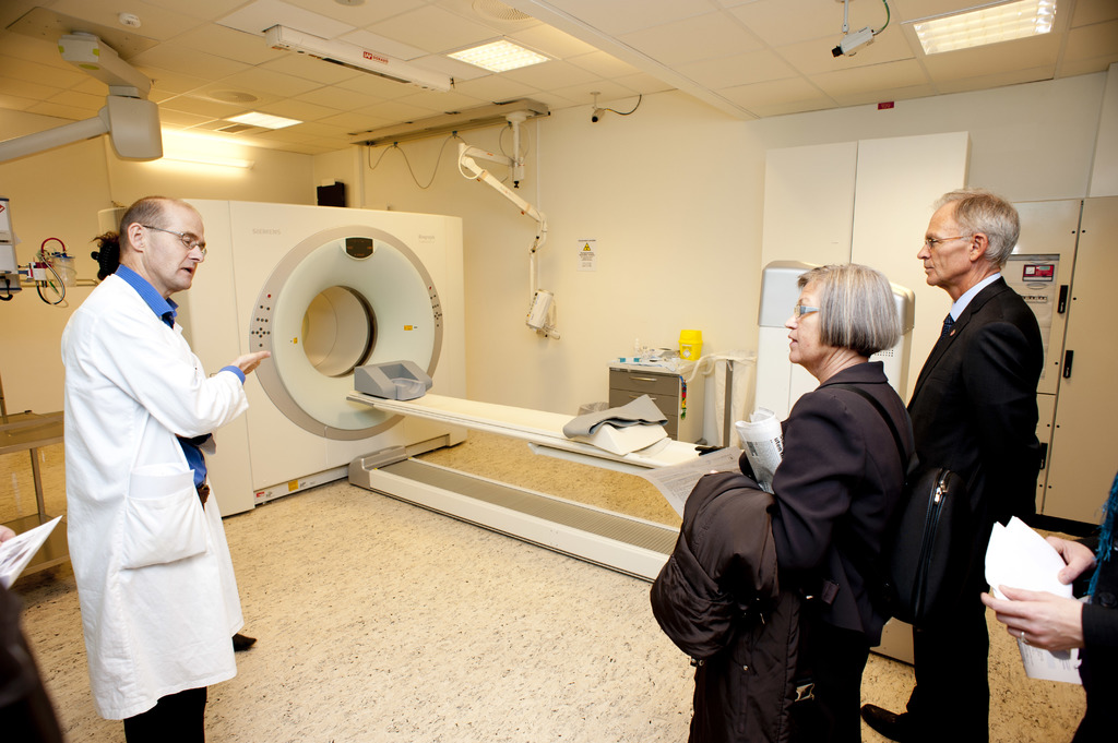 Ein Arzt in weißer Uniform erklärt eine CT-Scan-Maschine zwei Menschen in schwarzen Anzügen, die aufmerksam zuhören.