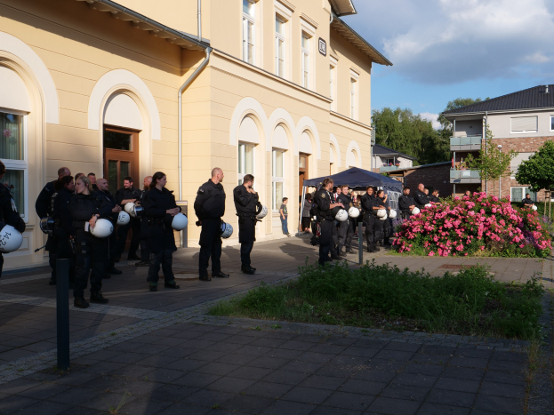Gruppe von Polizisten im Freien mit Helmen in der Hand vor einem Gebäude mit einem Zelt in der Nähe, umgeben von Grün und einem bewölkten Himmel.