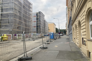 Stadtstraße mit Gebäuden, Fahrzeugen, Straßeninfrastruktur und einer Baustelle mit Gerüst im Vordergrund.
