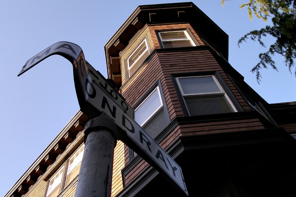 Ein Pfahl mit Text vorne, ein Gebäude in der Mitte und Blätter rechts.