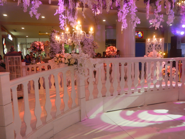 Weiße Reling mit Blumen und Kronleuchtern geschmückt über einer Hochzeitsfeier, mit Menschen drumherum, Säulen im Hintergrund, beleuchtet von Lichtern und einem Bildschirm auf der rechten Seite.
