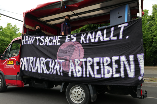 Roter Lkw mit einer Fahne fährt eine von Bäumen gesäumte Straße unter einem klaren blauen Himmel entlang, zwei maskierte Personen sind im Inneren sichtbar.