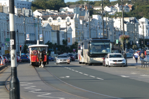 Vollständige Stadtstraße mit hohen Gebäuden, mehreren Fahrzeugen, Fußgängern auf dem Gehweg, Verkehrsampeln, Laternen, Informationsschildern, Bäumen und einem klaren blauen Himmel.