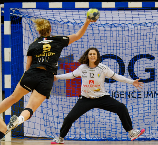Zwei Frauen in Sportkleidung beim Handballspielen auf einem Platz mit einem Tor und einer Banner im Hintergrund.