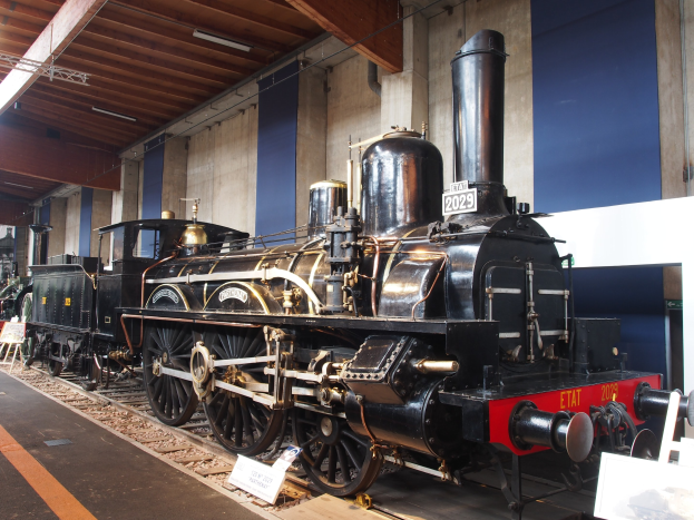 Eine große, alte Dampflokomotive auf Eisenbahnschienen in einem Museum ausgestellt, umgeben von Informationsschildern, Ständen und anderen Ausstellungsstücken, mit einer Wand und Deckenbeleuchtung im Hintergrund.