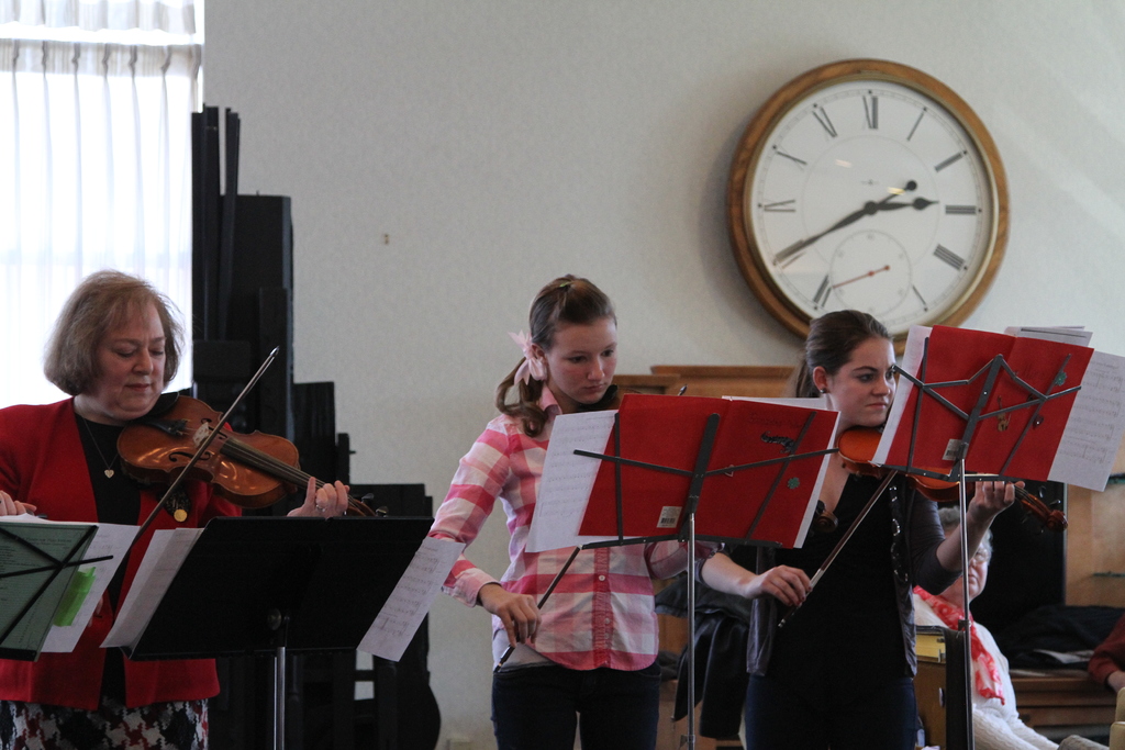 Drei Frauen, die Geigen spielen, stehen auf der Bühne vor Notenpulten, während eine Gruppe sitzender Frauen im Vordergrund zuhört; eine Uhr hängt an der weißen Wand im Hintergrund, mit einem weißen Vorhang in der linken Ecke.