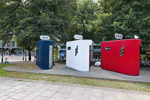 Drei mobile Toiletten auf einer grasbewachsenen Fläche neben einer Straße mit Gebäuden und einem klaren blauen Himmel im Hintergrund.
