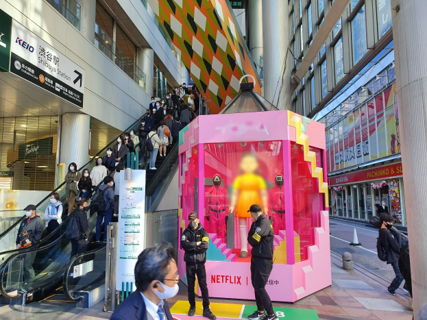Eine Gruppe von Menschen steht vor einem Gebäude mit Glaswänden und Texttafeln, einige tragen Masken, während eine Rolltreppe mit Menschen daneben fährt; ein pinker Kasten in der Mitte zeigt Schaufensterpuppen und trägt den Text "Netflix's new Netflix booth in Tokyo."