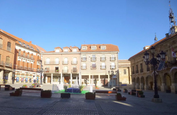 Ein Stadtplatz mit einem zentralen Brunnen, umgeben von Bänken, Topfpflanzen, Straßenlaternen und -pfosten, flankiert von Gebäuden und einem Uhrenturm unter einem klaren blauen Himmel.