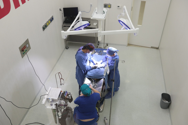 Chirurgen führen einen Eingriff an einem Patienten in einem Operationssaal durch, mit medizinischer Ausrüstung an der Wand links und einem Objekt auf dem Boden rechts.