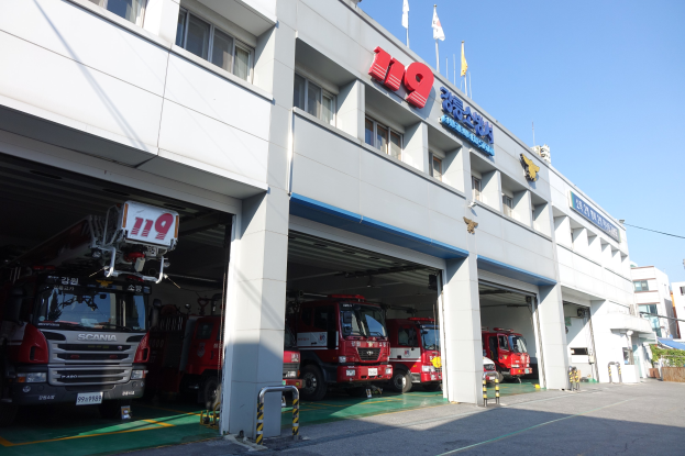 Feuerwache mit mehreren Feuerwehrautos vor der Tür, mit Fenstern, Säulen, Namensschildern und Flaggen mit Stangen im Vordergrund, vor anderen Gebäuden unter einem klaren blauen Himmel.