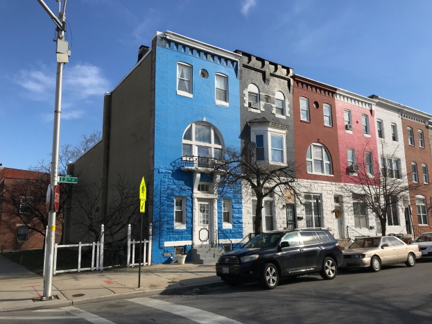 Eine Reihe bunter Häuser säumt eine Straßenkreuzung mit geparkten Autos, Bäumen, Strommasten, einem Zaun und einem bewölkten Himmel im Hintergrund.