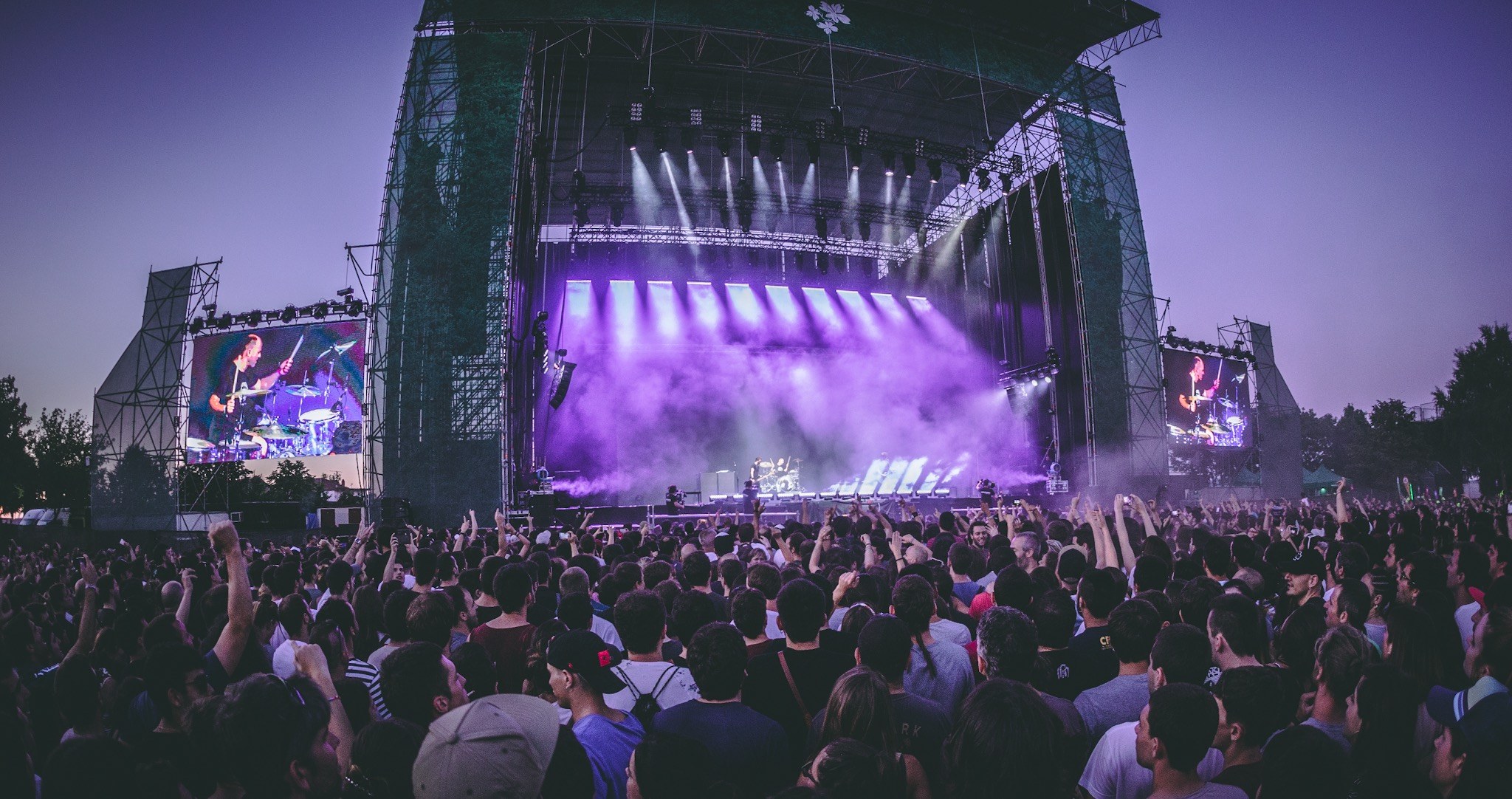 Eine große Menschenmenge auf einem Musikfestival schaut Künstlern auf einer hell erleuchteten Bühne mit Instrumenten und Bildschirmen zu, vor einem Gebäude, Bäumen und einem klaren blauen Himmel.