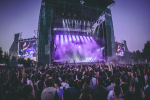 Eine große Menschenmenge auf einem Musikfestival schaut Künstlern auf einer hell erleuchteten Bühne mit Instrumenten und Bildschirmen zu, vor einem Gebäude, Bäumen und einem klaren blauen Himmel.