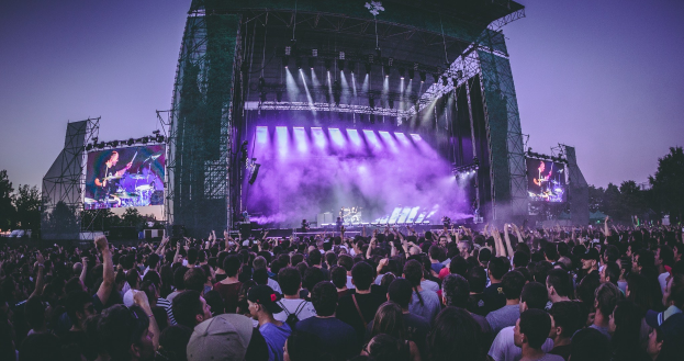 Eine große Menschenmenge auf einem Musikfestival schaut Künstlern auf einer hell erleuchteten Bühne mit Instrumenten und Bildschirmen zu, vor einem Gebäude, Bäumen und einem klaren blauen Himmel.