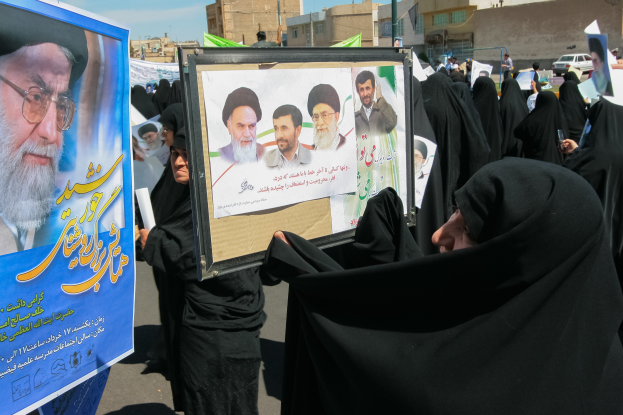 Eine Gruppe von Frauen in schwarzen Abayas hält Plakate mit Bildern von Ayatollah Ali Khamenei hoch, im Hintergrund sind Gebäude, Pfähle, Fahrzeuge und ein klarer blauer Himmel zu sehen.