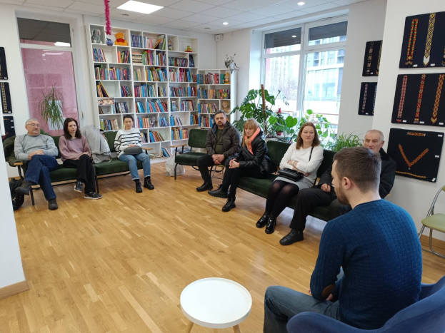 Gruppe von Menschen an einem Tisch in einem Raum mit Bücherregalen, Pflanzen und Deckenleuchten, die sich während eines Workshops zur Gesundheit und Wellness in Berlin, Deutschland, unterhalten.